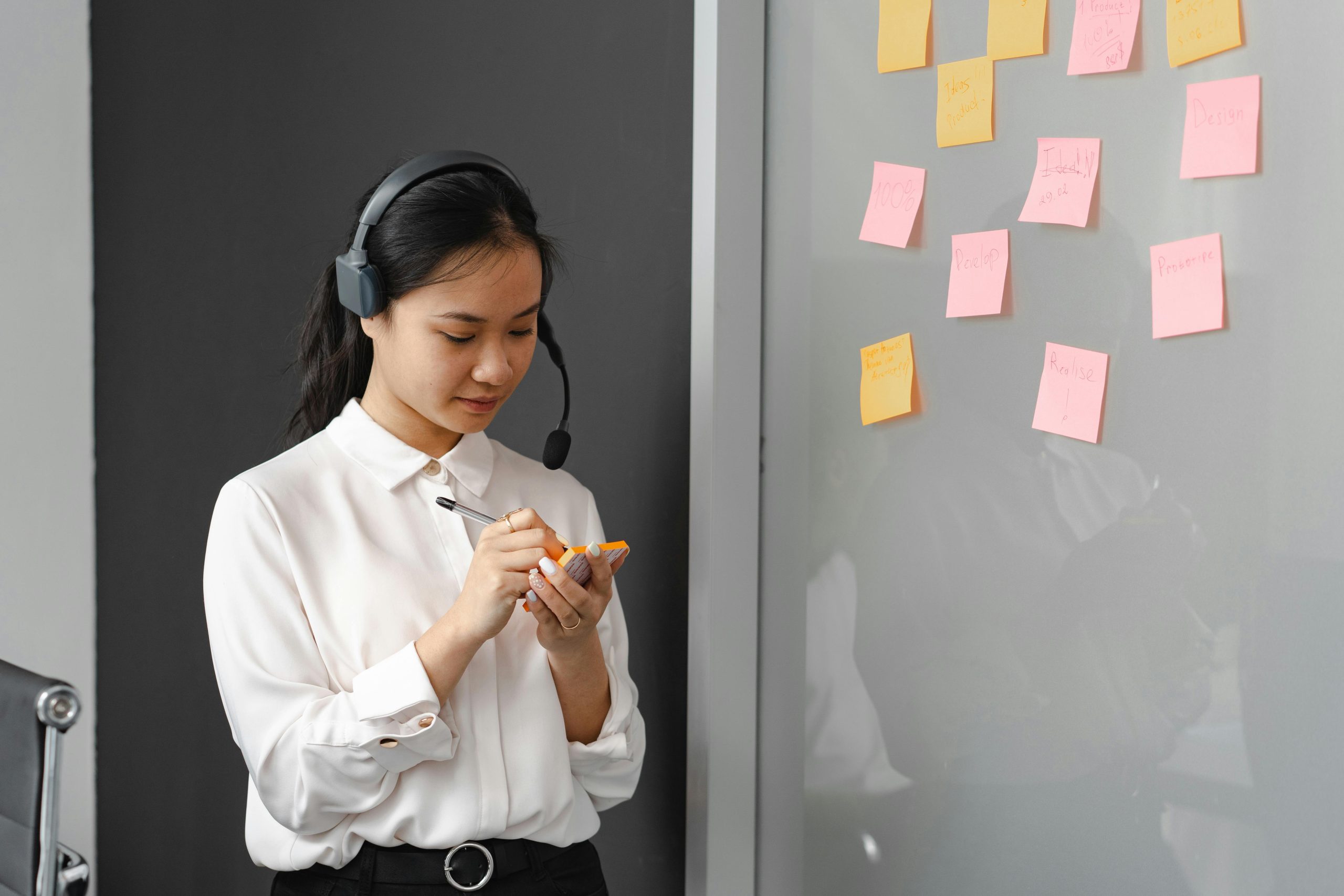 A woman in a call center writing on sticky notes with a headset indoors.